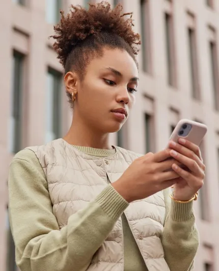 Vertical shot of curly haired woman uses mobile phone browses websites spends free time on social networks wears casual clothes stands against modern city building technology and lifestyle